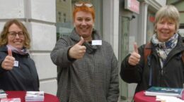 Drei Frauen auf dem Bürgersteig in der Einkaufsstraße hinter Stehtischen halten lächelnd Daumen hoch.