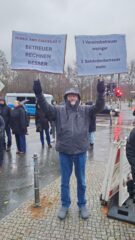Ein Mann steht im Regen und hält zwei Plakate hoch. Schriftzug Betreuer rechnen besser und 1 Vereinsbetreuer weniger gleich 2 Behördenbetreuer mehr