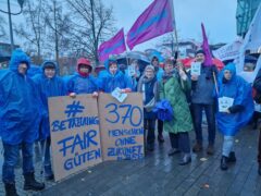 Protestierende im Regen halten großen Pappschilder mit dem Schriftzug #BetreuungFairgüten und 370 Menschen ohne Zukunft in BRB. Flaggen der Diakonie